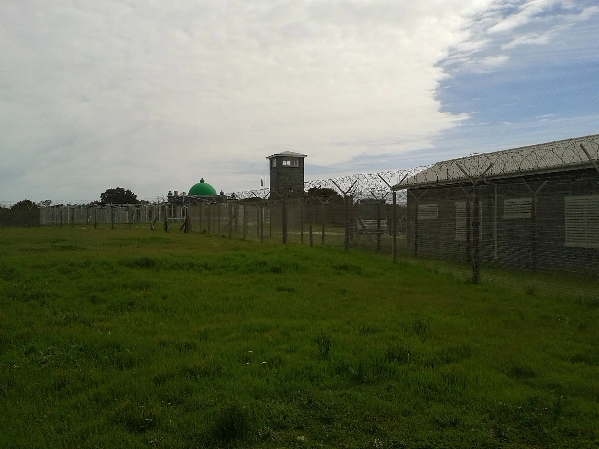 Barbed wire and a watchtower around the prison, with the green dome of the Moturu Kramat behind